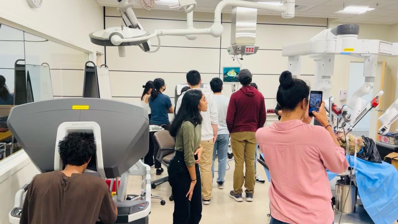 A group of people in a medical or research lab interacting with robotic surgical equipment. One person in the foreground is seated at a Da Vinci surgical console, while another is taking a photo with a smartphone. Several individuals in the background are gathered around a screen displaying a 3D model, while others are observing robotic arms covered in blue surgical drapes. The room is well-lit, with advanced medical technology and monitors mounted on the ceiling.