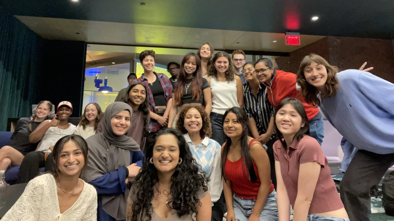 A large group of smiling young adults pose together indoors in a well-lit lounge or common area with dark ceilings and modern decor. The group consists of approximately twenty people, some seated and others standing closely behind them. They appear to be part of a class or program, with several individuals leaning in cheerfully toward the camera. The atmosphere is friendly and relaxed, and the space behind them includes glass walls, soft lighting, and a mix of seating areas.
