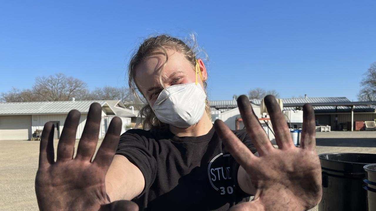 A person wearing a white mask and black T-shirt stands outdoors on a sunny day, holding up both hands, covered in black dirt and kelp, toward the camera. The person has light-colored hair pulled back and is standing in a gravel lot with metal buildings and trees in the background. A utility cart is partially visible on the right side of the image