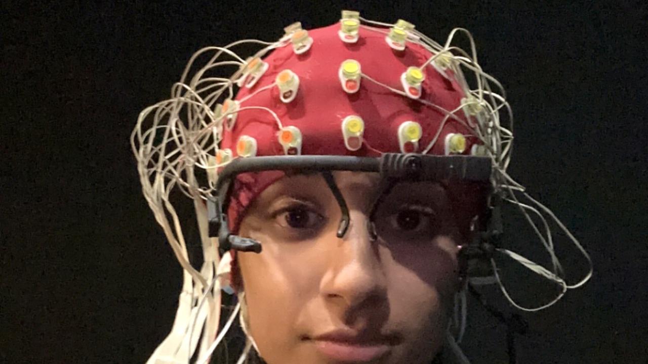 A person wearing a white EEG cap covered with numerous electrodes and wires sits facing the camera. Additional sensors are attached to their face, including one on the forehead with a circular black-and-white target design and another near the cheek. The person is wearing a light-colored sweatshirt and is seated against a dark background, likely in a research or clinical setting.