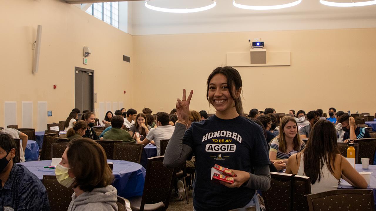 A student wearing a T-shirt with the words Honored to be an Aggie gestures a peace sign while standing a room filled with other students at round tables.