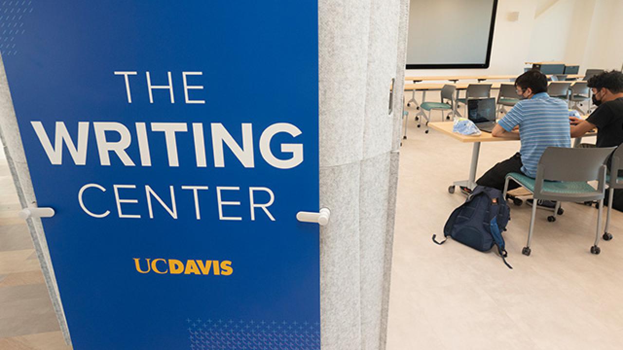 Blue sign on a pillar that says the Writing Center UC Davis. Behind the pillar, two students are working on laptops at a table.