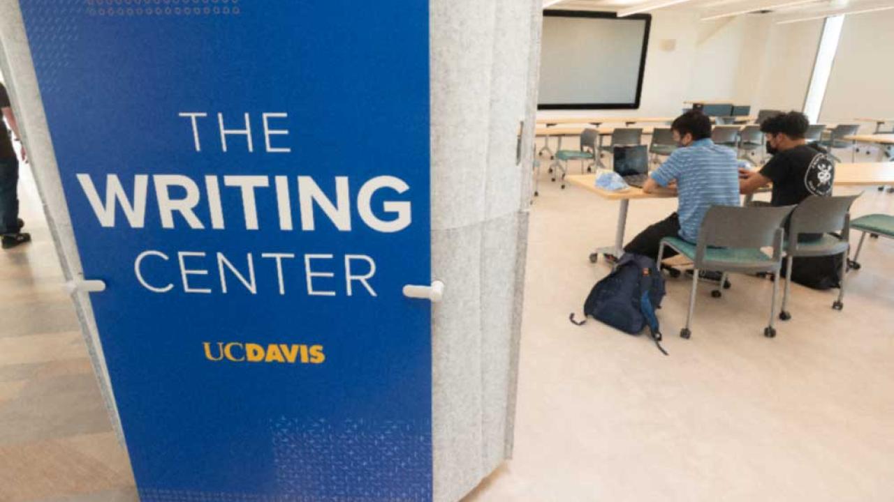 The photo features a 8-foot-tall blue sign attached toa wall that says Writing Center. Beside the wall is an opening into a classroom in the Writing Center. The viewer can see  several long work tables with two people working on laptops seated next to each other at the nearest table. 