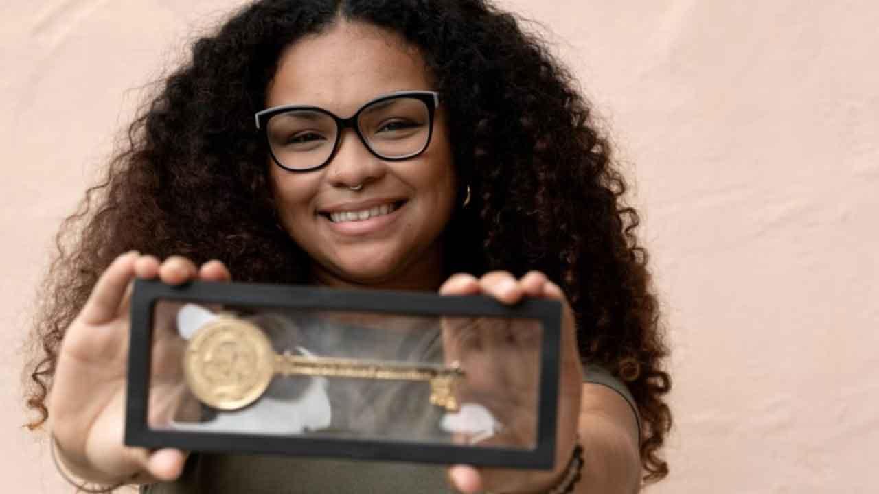 A smiling person with curly hair and glasses holds a rectangular display case toward the camera, showcasing a large gold key with an emblem on one end. The person wears a nose ring and is standing in front of a light-colored wall. The focus is on the key.