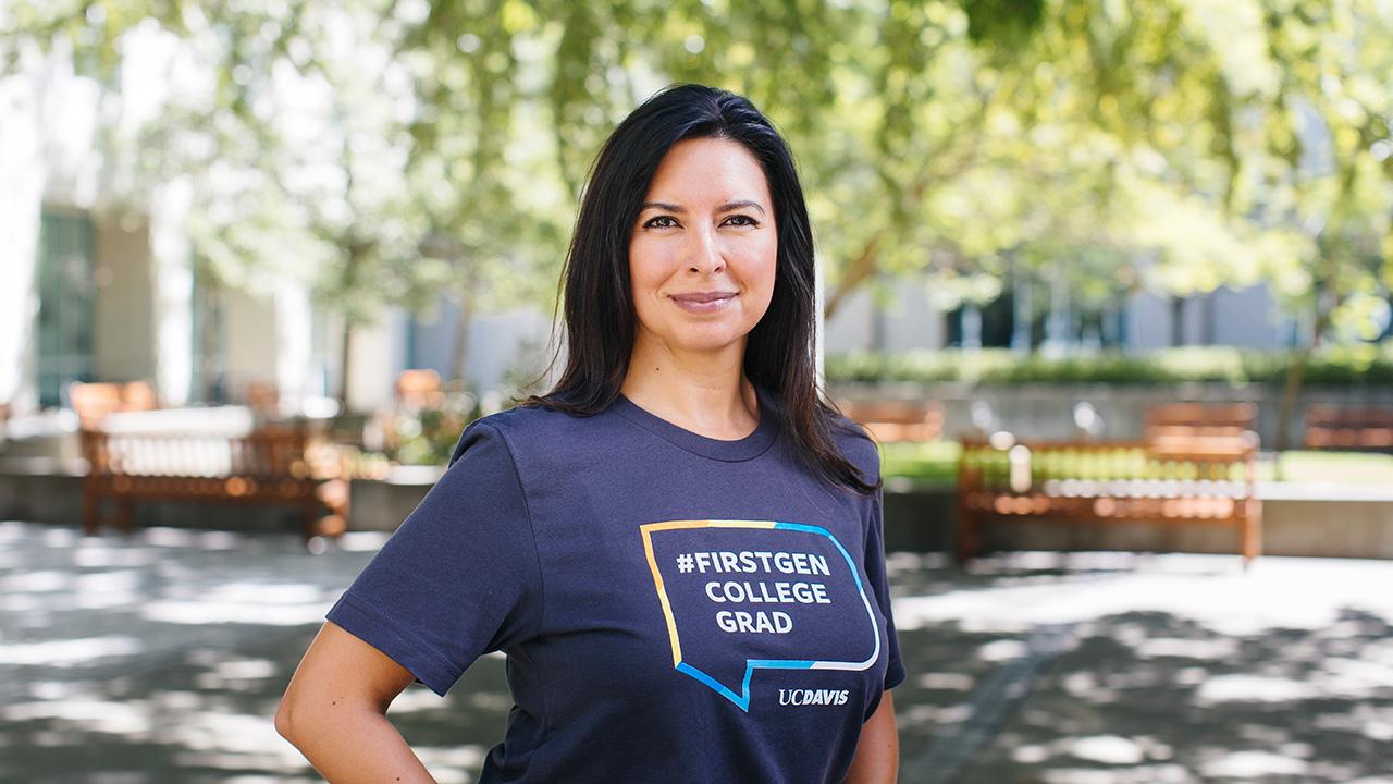 Jeanette B. Ruiz stands with a hand on her hip in the middle of Shields Library courtyard. She is wearing a blue T-shirt with a colorful graphic that says #firstgen college grad UC Davis.