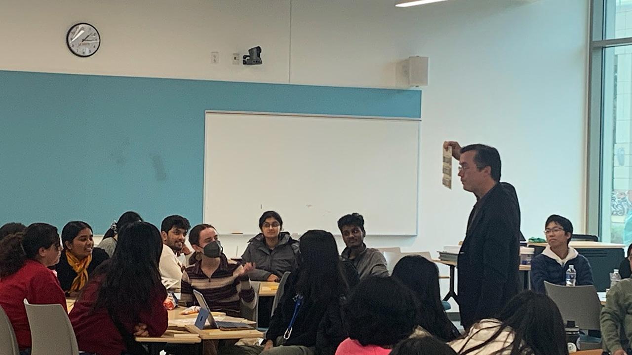 A professor, standing in a large room surrounded by seated students, holds up a paper-sized board for students to see.