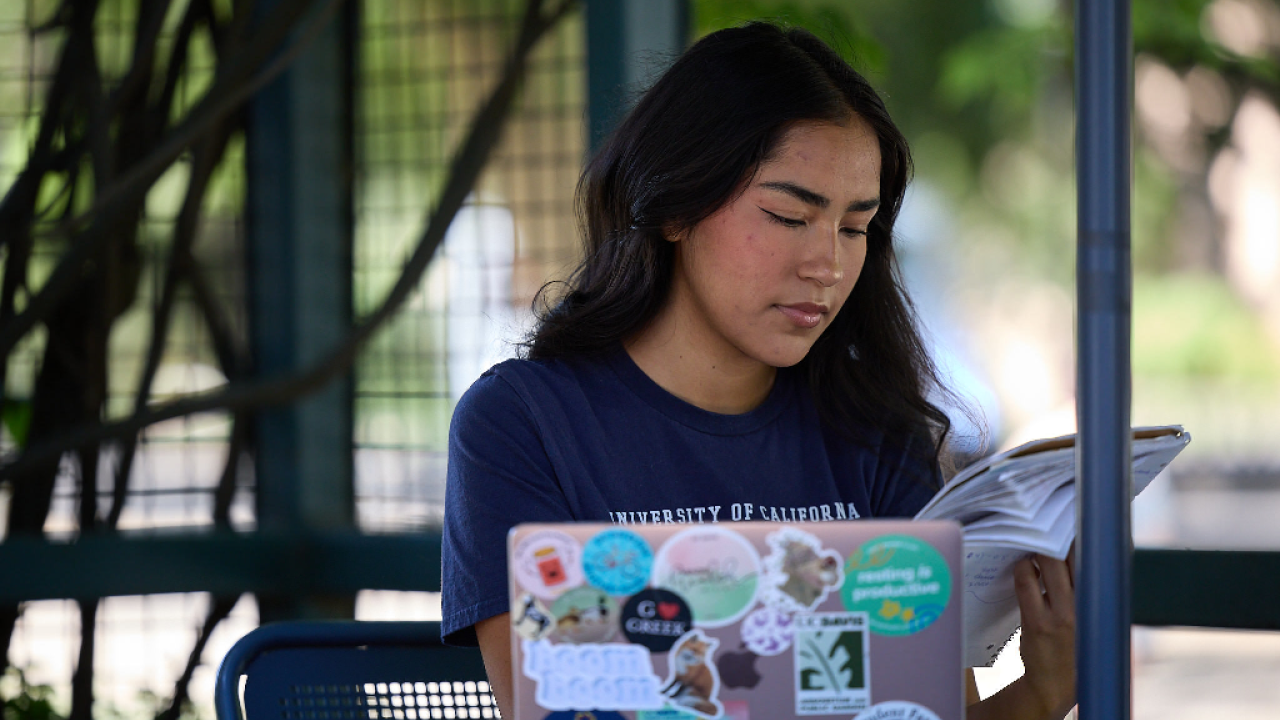  A student seated at an outdoor table with her laptop in front of her while she looks through a notebook.