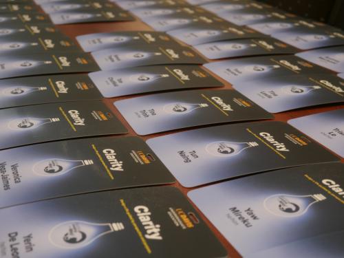 A table covered with neatly arranged name badges for an event. The badges are dark blue with a gradient effect and feature the word 'Clarity' along with a lightbulb graphic. Each badge displays a participant's name and role. The perspective of the image shows the badges extending into the distance, creating a sense of depth.