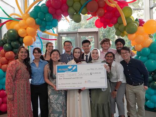 Team of 11 student entrepreneurs holds up a giant check for $10,000 while posing under a colorful balloon arch. 