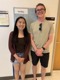 Two smiling individuals stand side by side in a hallway in front of a bulletin board labeled 'Department of Sociology.' The person on the left is wearing a maroon cardigan, dark top, and denim shorts. The person on the right is wearing glasses, a light green T-shirt, black shorts, and a watch, with sunglasses hanging from their shirt.