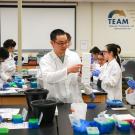 Multiple students sitting in a laboratory classroom with some students watching another student point at a lab tool.