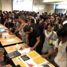 Large group of students gathered around multiple tables set up in rows. 