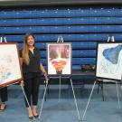 A woman with long brown hair, wearing a black outfit, stands smiling in front of three framed paintings displayed on easels in an indoor exhibition space. The artwork on the left features an abstract composition with splashes of color and floral elements. The middle piece portrays a surreal landscape with layered, colorful hills inside a floating shape against a night sky with trees. The artwork on the right consists of a dark blue abstract form with splattered paint on a white background.