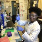 A person wearing a white lab coat, safety goggles, and blue gloves uses a pipette at a lab workstation. The workstation is filled with colorful lab supplies including microcentrifuge tubes, racks, pipette tips, and various containers. The individual is smiling slightly and appears to be in the middle of an experiment.
