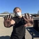 A person wearing a white mask and black T-shirt stands outdoors on a sunny day, holding up both hands, covered in black dirt and kelp, toward the camera. The person has light-colored hair pulled back and is standing in a gravel lot with metal buildings and trees in the background. A utility cart is partially visible on the right side of the image