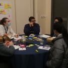 A group of five people sitting around a table engaged in discussion during a workshop session. The table is covered with a blue tablecloth and scattered with sticky notes, pens, paper and snacks. One participant is wearing a face mask and taking notes, while others listen and converse. A large sheet of paper with colorful sticky notes is attached to the wall in the background.