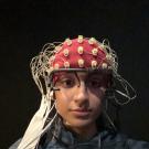 A person wearing a white EEG cap covered with numerous electrodes and wires sits facing the camera. Additional sensors are attached to their face, including one on the forehead with a circular black-and-white target design and another near the cheek. The person is wearing a light-colored sweatshirt and is seated against a dark background, likely in a research or clinical setting.