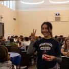 A student wearing a T-shirt with the words Honored to be an Aggie gestures a peace sign while standing a room filled with other students at round tables.