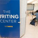 Blue sign on a pillar that says the Writing Center UC Davis. Behind the pillar, two students are working on laptops at a table.