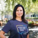 Jeanette B. Ruiz stands with a hand on her hip in the middle of Shields Library courtyard. She is wearing a blue T-shirt with a colorful graphic that says #firstgen college grad UC Davis.