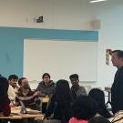 A professor, standing in a large room surrounded by seated students, holds up a paper-sized board for students to see. 