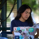  A student seated at an outdoor table with her laptop in front of her while she looks through a notebook.
